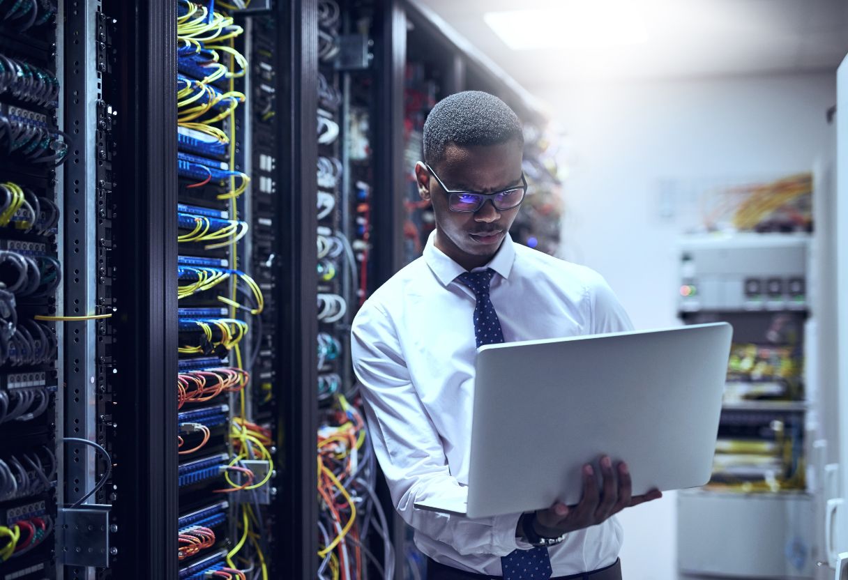 A professional man in a white shirt and tie holds a laptop in a server room filled with colorful data cables and racks, searching for server jobs near me to advance his career in the tech industry.